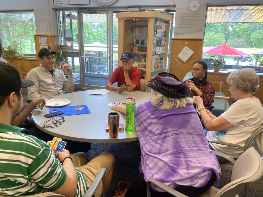 A group of people sitting around a round table inside a building with large windows in the background looking out onto a patio scene. There are playing cards from the game UNO! in several people's hands. One person in a purple shirt and black hat is reaching towards the centre of the table towards a stack of cards piled up in the middle of the table.