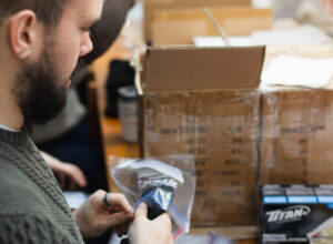 A person with their back to the camera holds an item that they are putting into a clear plastic bag. Next to them are some boxes labeled to be sent to another location. In the background, several people seem to be doing a similar task.